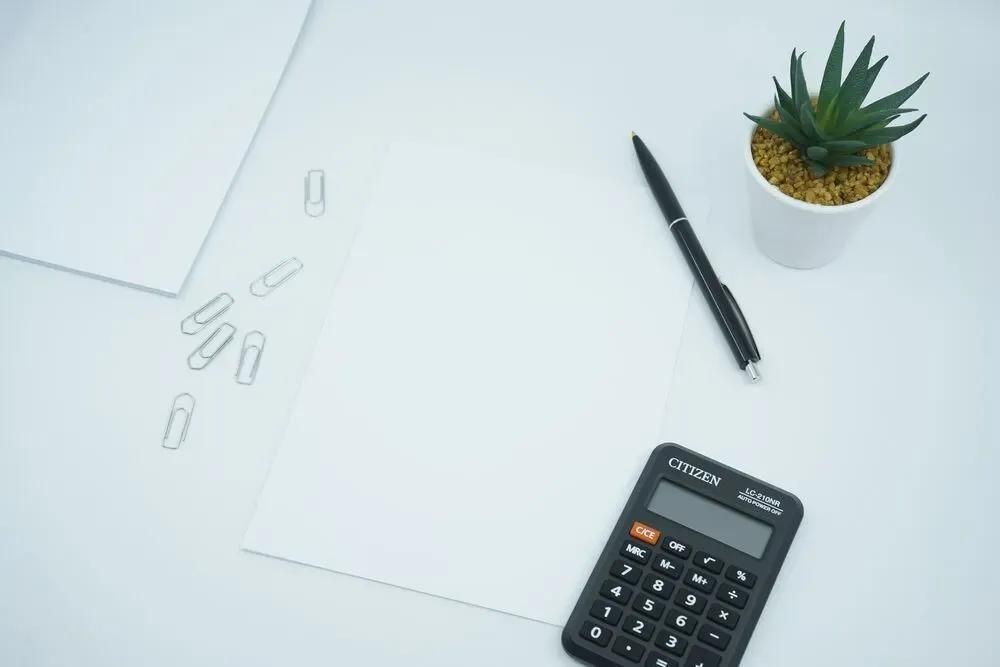 Taschenrechner, weisses Blatt, schwarzer Stift, Büroklammern und Pflanze auf Büro